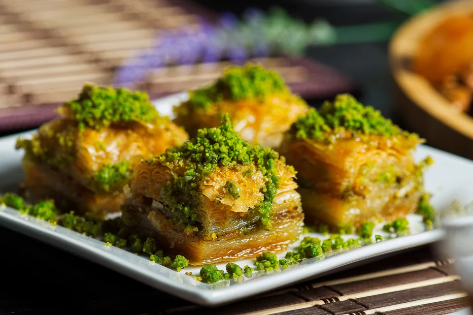 BookNoFees: Halimbawa ng larawan: Apat na piraso ng baklava na may patong-patong na layer at tinadtad na pistachio sa ibabaw, sa isang puting plato